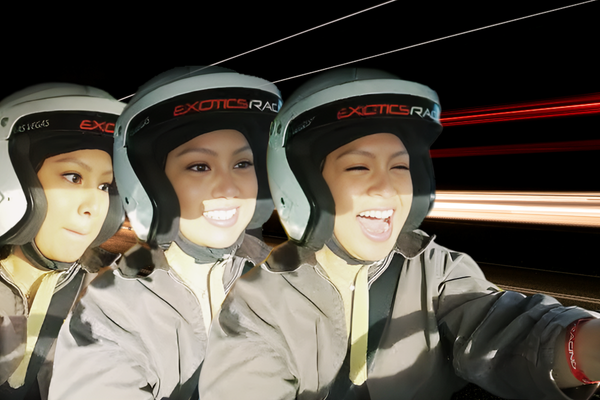 Three women wearing white racing helmets and gray driving suits sit side-by-side in a high-speed exotic car experience, smiling and reacting with excitement against a blurred light-trail background.