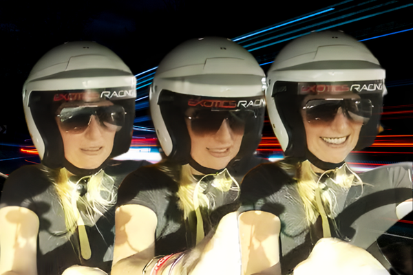 Three women wearing black “Exotics Racing” helmets and sunglasses lean forward in concentration while driving, illuminated by colorful motion light streaks suggesting high speed.