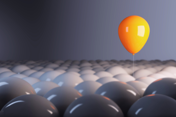 Bright orange balloon floating above a field of gray spheres, representing standing out, uniqueness, and differentiation in a competitive market.