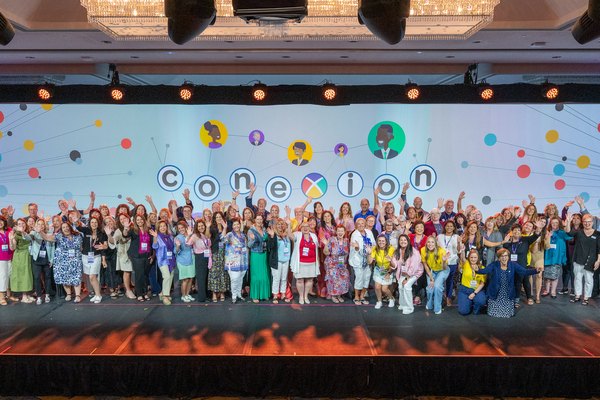 Nexion Travel Group advisors gather on stage in front of the colorful coneXion conference backdrop, waving and celebrating together